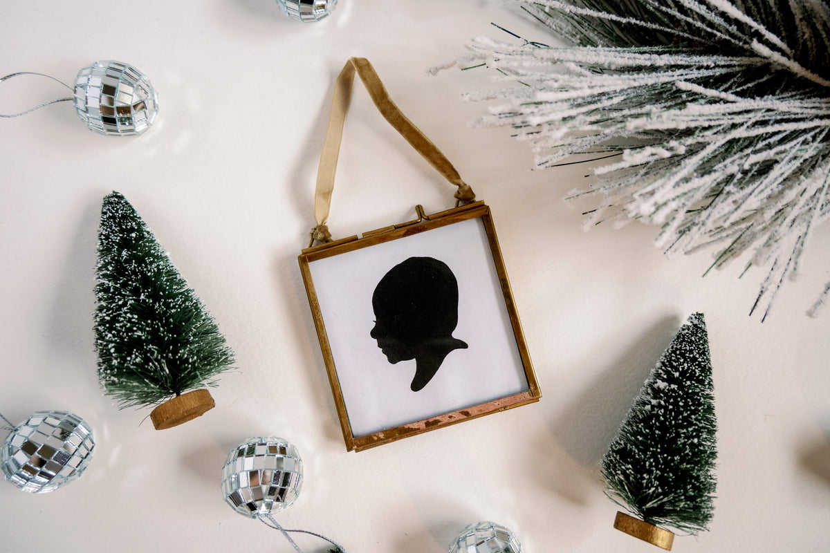 A square, gold-framed ornament featuring a black silhouette of a childs profile, surrounded by miniature evergreen trees and small disco ball ornaments on a white surface.