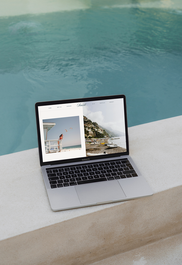 A laptop displaying a travel or photography website built with a Showit template rests on the edge of a light-colored poolside ledge, with clear blue pool water in the background.